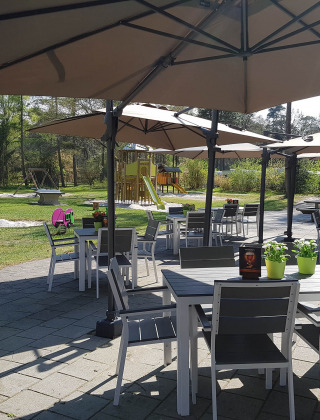 Zona de asientos al aire libre con mesas, sillas y sombrillas grandes, parque infantil al fondo en Drenthe, Países Bajos.