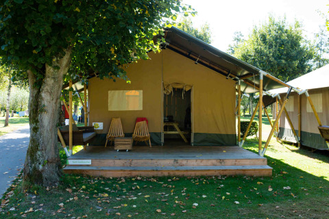Outdoor photo of a Safari tent lodge featuring a wooden porch, chairs, and peaceful green surroundings.