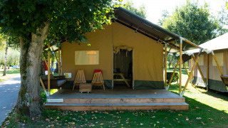 Outdoor photo of a Safari tent lodge featuring a wooden porch, chairs, and peaceful green surroundings.