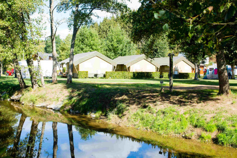 Safari tents at Camping Val d'Or in Luxembourg, seen by a small creek and surrounded by greenery and trees.