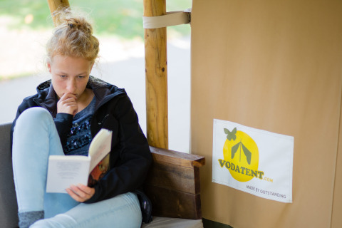 En pige læser en bog i en safaritelt-lodge på Camping Val d'Or i Luxembourg, ved siden af Vodatent-logoet.