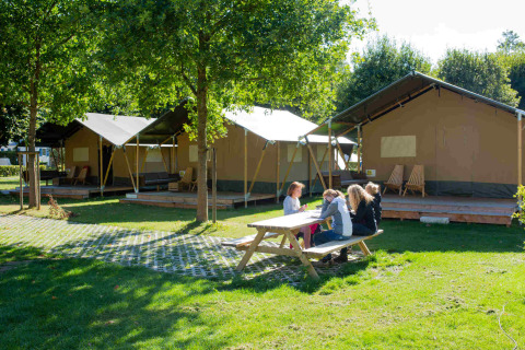Mesa de picnic al aire libre junto a tiendas safari en Camping Val d'Or, Luxemburgo, con personas sentadas.