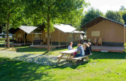 Mesa de picnic al aire libre junto a tiendas safari en Camping Val d'Or, Luxemburgo, con personas sentadas.
