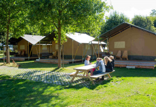 Mesa de picnic al aire libre junto a tiendas safari en Camping Val d'Or, Luxemburgo, con personas sentadas.