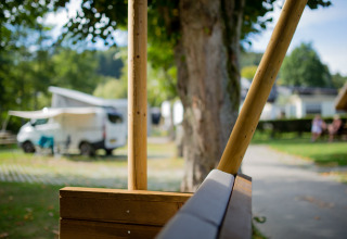 Vista desde un banco de madera hacia una caravana y vegetación en Camping Val d'Or, Luxemburgo.