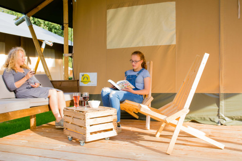 Dos personas relajándose frente a una tienda safari en Camping Val d'Or en Luxemburgo, tomando bebidas.