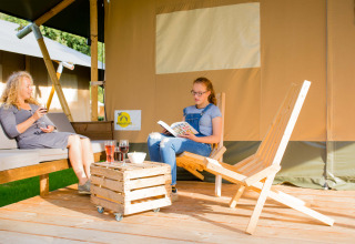 Dos personas relajándose frente a una tienda safari en Camping Val d'Or en Luxemburgo, tomando bebidas.