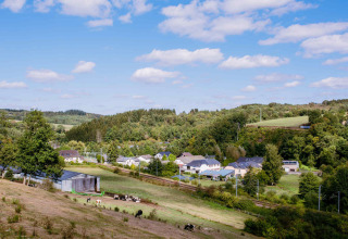 Scenic view of Camping Val d'Or in Wiltz, Luxembourg, with green fields, houses, cows, and rolling wooded hills.