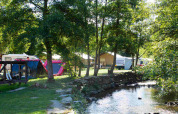 Campingplads ved flod med telte, campingvogne og træer i sollys på Camping Val d'Or i Wiltz, Luxembourg.
