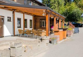 Udendørs terrasse med borde og stole ved Camping Val d'Or i Wiltz, Luxembourg, omgivet af grønne træer.