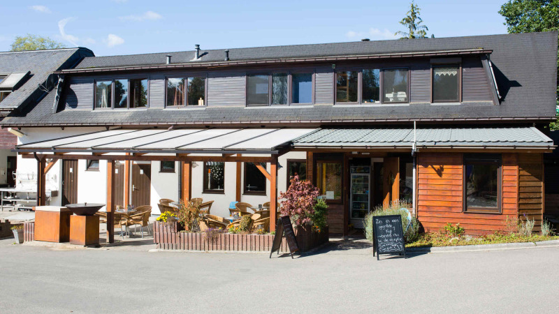 Außenansicht des Hauptgebäudes von Camping Val d'Or mit Terrasse in Wiltz, Luxemburg, bei sonnigem Wetter.