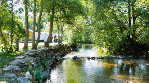 Sunny campsite by a peaceful stream in wooded surroundings, with tents and caravans at Camping Val d'Or, Luxembourg.