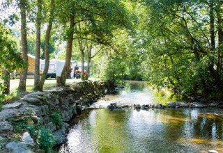 Solrig campingplads ved en fredelig å i skovområde, med telte og campingvogne på Camping Val d'Or, Luxembourg.