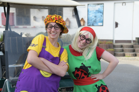 Dos personas alegres con disfraces coloridos y gafas posan frente a un carrito en Camping Val d'Or, Luxemburgo.