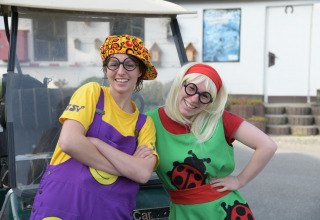 Dos personas alegres con disfraces coloridos y gafas posan frente a un carrito en Camping Val d'Or, Luxemburgo.