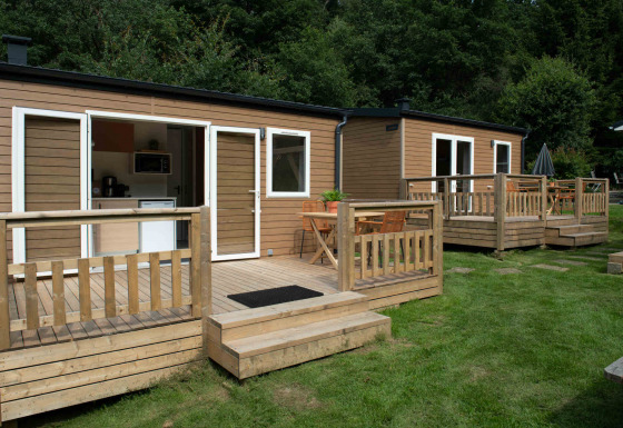 Twee kleine tiny houses met houten terrassen op gras, omgeven door bomen in een groene omgeving.