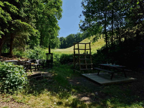 Udendørs siddeområde med borde og stole i frodig natur ved Boslodge um Bierg, Camping Val d'Or, Luxembourg.