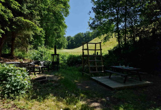 Udendørs siddeområde med borde og stole i frodig natur ved Boslodge um Bierg, Camping Val d'Or, Luxembourg.