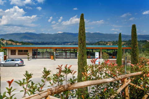 Parque vacacional en la Toscana, Italia, con edificio moderno, cipreses y montañas visibles a lo lejos bajo cielo azul.