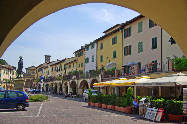 Foto van een schilderachtig Toscaans dorpsplein in Italië met restaurants, beelden en geparkeerde auto's.