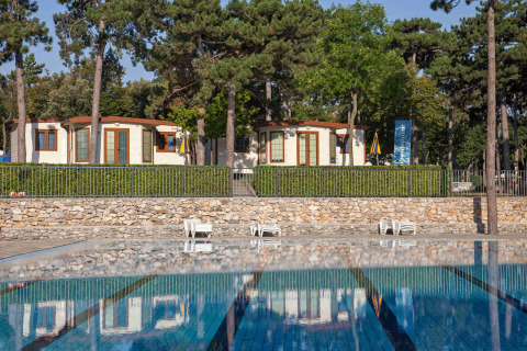 Vista de la piscina y bungalows rodeados de pinos en Camping Village Mare Pineta, Friuli-Venezia Giulia, Italia.