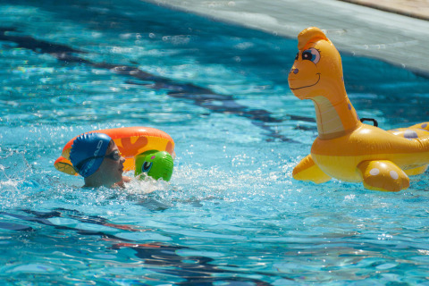 Bambino nuota con ciambella accanto a un dinosauro gonfiabile in piscina al Camping Village Mare Pineta, Italia.