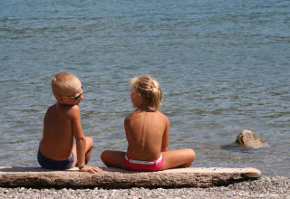 Two children sit on a log by the water at Camping Village Mare Pineta in Friuli-Venezia Giulia, Italy.
