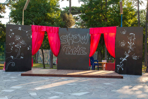 Palco all’aperto con tende rosse, pannelli neri decorati con fiori e scritta 'SHOW TIME' al centro.