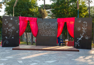 Palco all’aperto con tende rosse, pannelli neri decorati con fiori e scritta 'SHOW TIME' al centro.