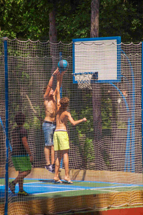 Niños juegan baloncesto en una cancha exterior con red en Camping Village Mare Pineta, Friuli-Venezia Giulia, Italia.