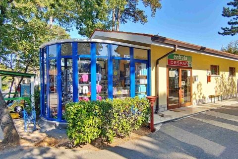 Ingang van de Despar supermarkt bij Camping Village Mare Pineta in Friuli-Venezia Giulia, Italië, omgeven door groen.