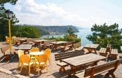Udendørs terrasse med træborde og gule stole, udsigt over havet og klipper, Mare Pineta, Italien.