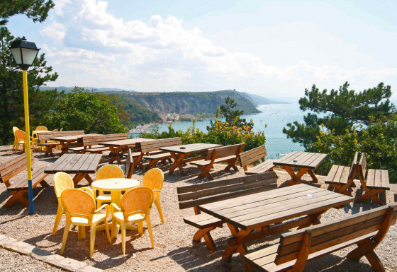 Udendørs terrasse med træborde og gule stole, udsigt over havet og klipper, Mare Pineta, Italien.