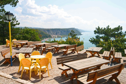 Udendørs terrasse med træborde og gule stole, udsigt over havet og klipper, Mare Pineta, Italien.