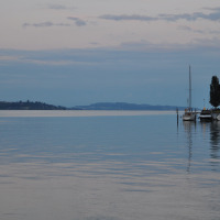Vista de un lago tranquilo con veleros atracados junto a un parque vacacional con glamping al atardecer.