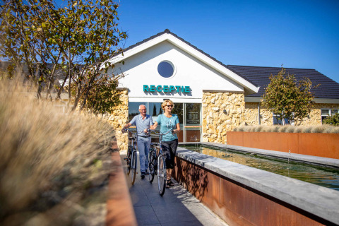Dos personas con bicicletas junto a la recepción del Resort Mooi Bemelen en Limburg, Países Bajos, en un día soleado.