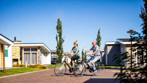 Ældre par cykler på ferieparken Resort Mooi Bemelen i Limburg, Holland, under en solrig himmel.