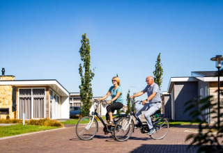 Ældre par cykler på ferieparken Resort Mooi Bemelen i Limburg, Holland, under en solrig himmel.