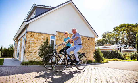 Un couple âgé fait du vélo devant un cottage en pierre au Resort Mooi Bemelen, Limburg, Pays-Bas, en été.