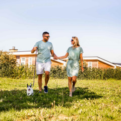Pareja paseando con perro en Resort Mooi Bemelen, parque vacacional en Limburg, Países Bajos, día soleado.
