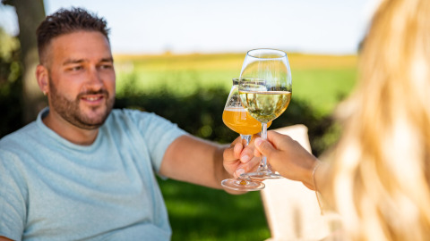 Twee mensen proosten met wijn en bier in de buitenlucht bij Resort Mooi Bemelen, een vakantiepark in Limburg, Nederland.
