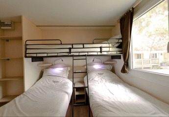 Glamping accommodation room with two single beds, overhead bunk, shelves and a large window, cozy setup.