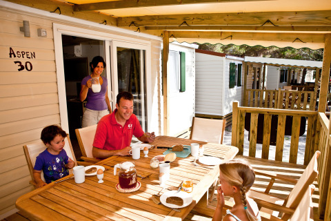 Una familia desayuna junta en la terraza de una cabaña, rodeada de casas móviles y un ambiente soleado.