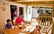 Una familia desayuna junta en la terraza de una cabaña, rodeada de casas móviles y un ambiente soleado.