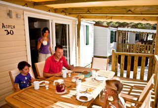 Una familia desayuna junta en la terraza de una cabaña, rodeada de casas móviles y un ambiente soleado.