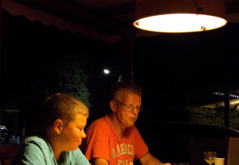 Two people sit at a table under a lamp in a lodge at night, playing a dice game and talking quietly.