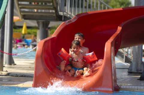 Two children laugh and enjoy a red water slide by the pool at Camping Pra'delle Torri, Veneto, Italy.