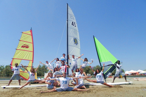 Young adults cheerfully posing in front of colorful sailboats on the beach at Camping Pra'delle Torri, Italy.