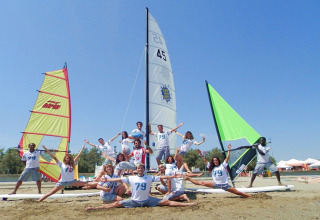 Young adults cheerfully posing in front of colorful sailboats on the beach at Camping Pra'delle Torri, Italy.