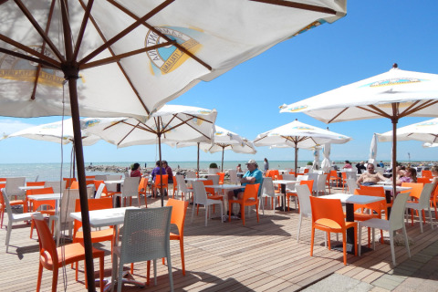 Zona exterior de cafetería con sillas naranjas y blancas, sombrillas y vista al mar en Camping Pra'delle Torri.